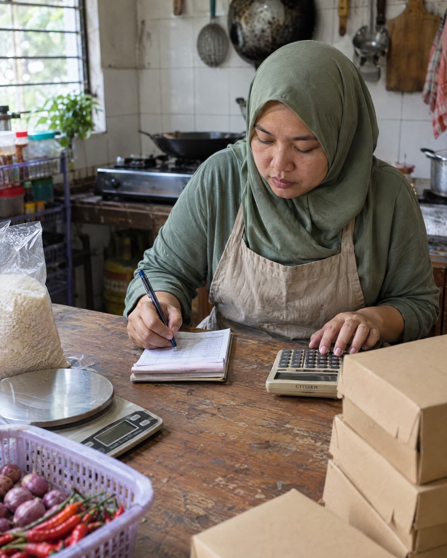 Pemilik usaha makanan rumahan mencatat modal di buku nota sambil memakai kalkulator